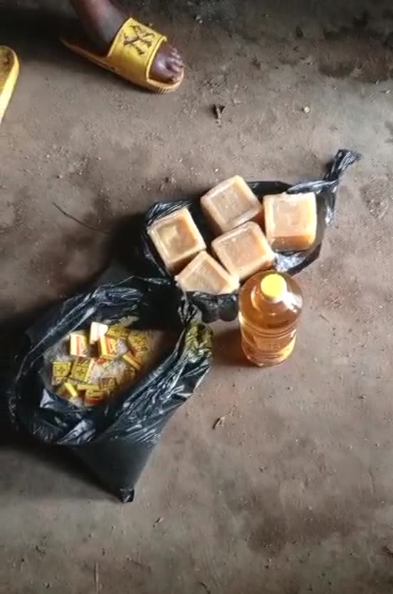 Bars of soap, a bottle of cooking oil and a bag of seasoning laid out on a swept earth floor, beside a sandal.